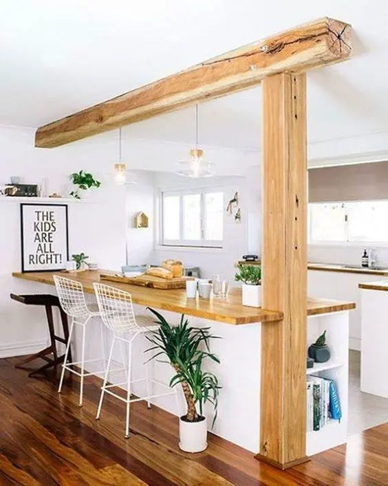Espacio abierto con barra desayunadora de madera sostenida por una viga, acompañada de sillas altas y toques naturales, generando un ambiente acogedor y armónico