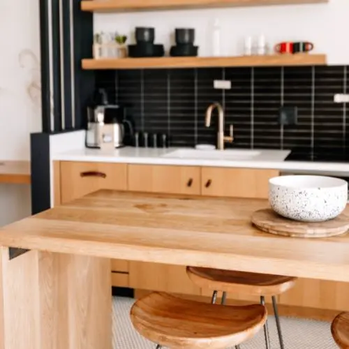 Cocina moderna con isla de madera, sillas redondas y fondo de azulejos negros, creando un ambiente cálido y funcional