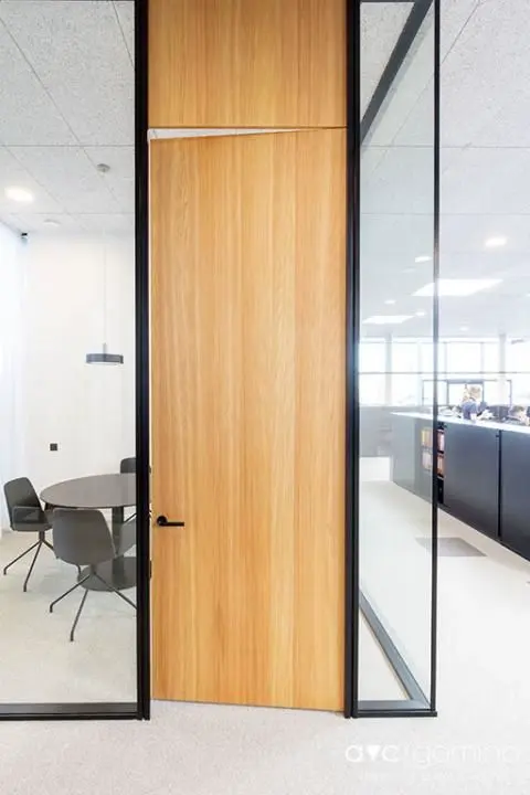 Puerta de madera de altura completa con marco de vidrio lateral, integrada en un entorno de oficina moderno y luminoso.