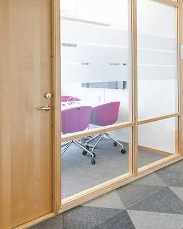 Puerta de madera con grandes paneles de vidrio en una sala de reuniones con sillas modernas y alfombra de oficina.
